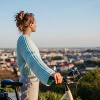 A person wearing a cozy sweater stands with her bicycle on a hill overlooking a city